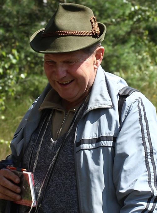 Ein lachender älterer Mann mit Jägerhut, Fernglas und einem Notizbuch in der Natur